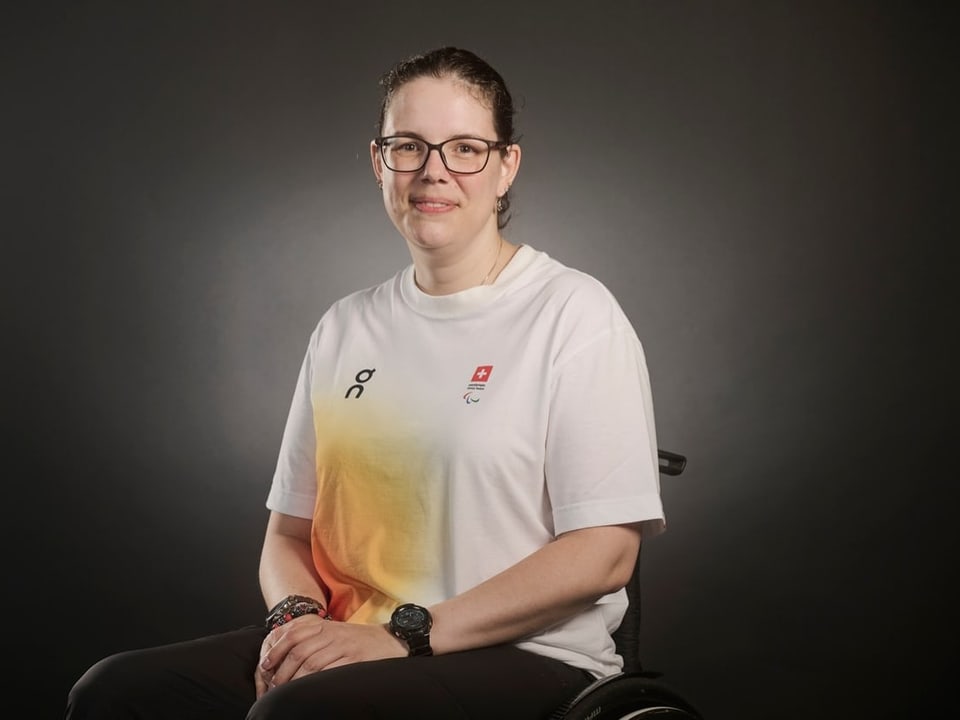 Frau im Rollstuhl mit Brille und weissem T-Shirt vor neutralem Hintergrund.