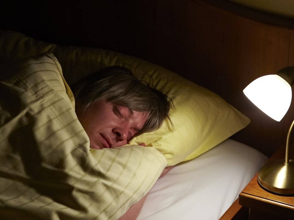 Person schläft im Bett neben eingeschalteter Lampe.