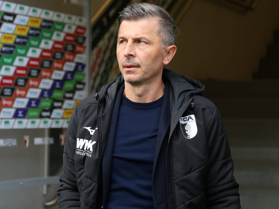 Fussballtrainer in schwarzer Jacke vor gesponserter Wand.