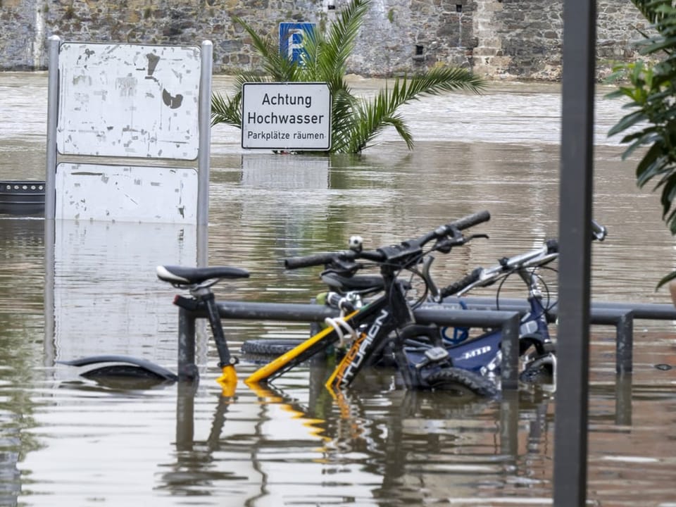 Überfluteter Parkplatz mit Fahrrädern und Warnschild für Hochwasser.