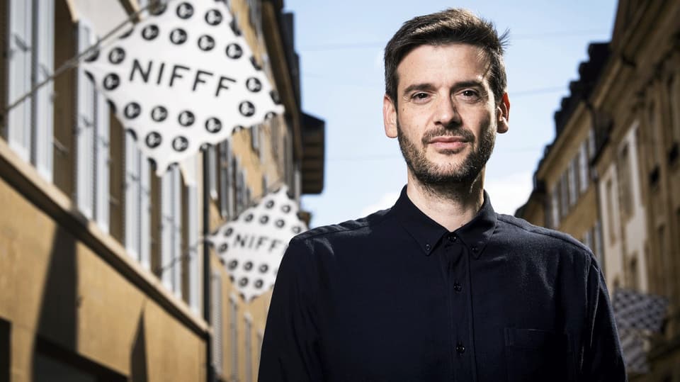 Ein Mann mit kurzen Haaren und Hemd steht in einer Gasse, an den Häuserfassaden hängen Fahnen mit dem Schriftzug NIFFF.