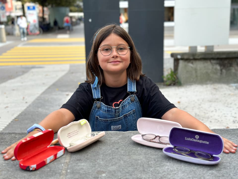 Ein Mädchen sitzt vor vier Brillenetuis mit verschiedenen Brillen darin. 