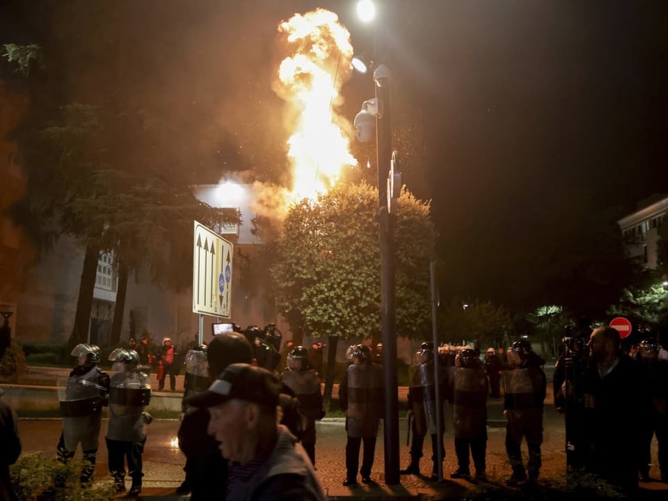 Nachtszene mit brennendem Gebäude und Polizei in Schutzkleidung.