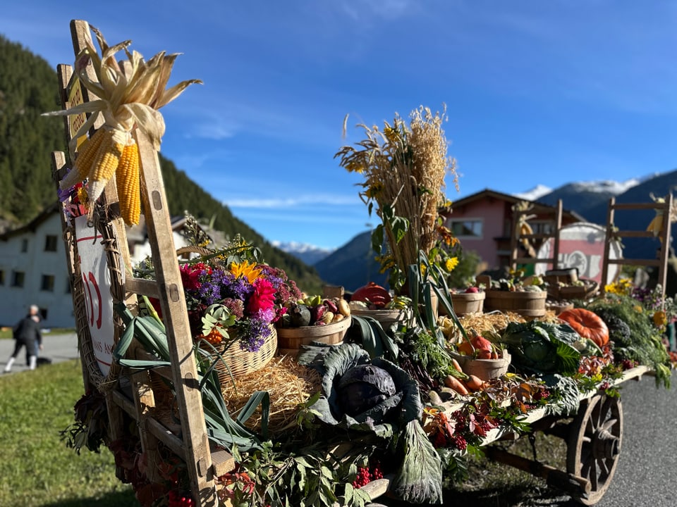 Dekorativer Erntewagen mit Gemüse und Blumen in alpiner Landschaft.