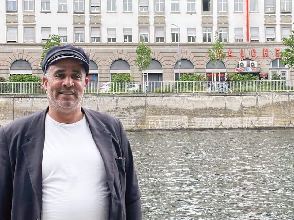 Ein Mann in weissem T-Shirt, grauem Blazer und grauer Schiebermütze steht vor dem Fluss Limmat in Zürich. 