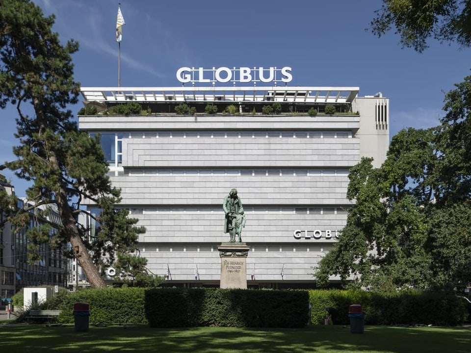 Gebäude mit Globus-Logo hinter Statue und Bäumen.