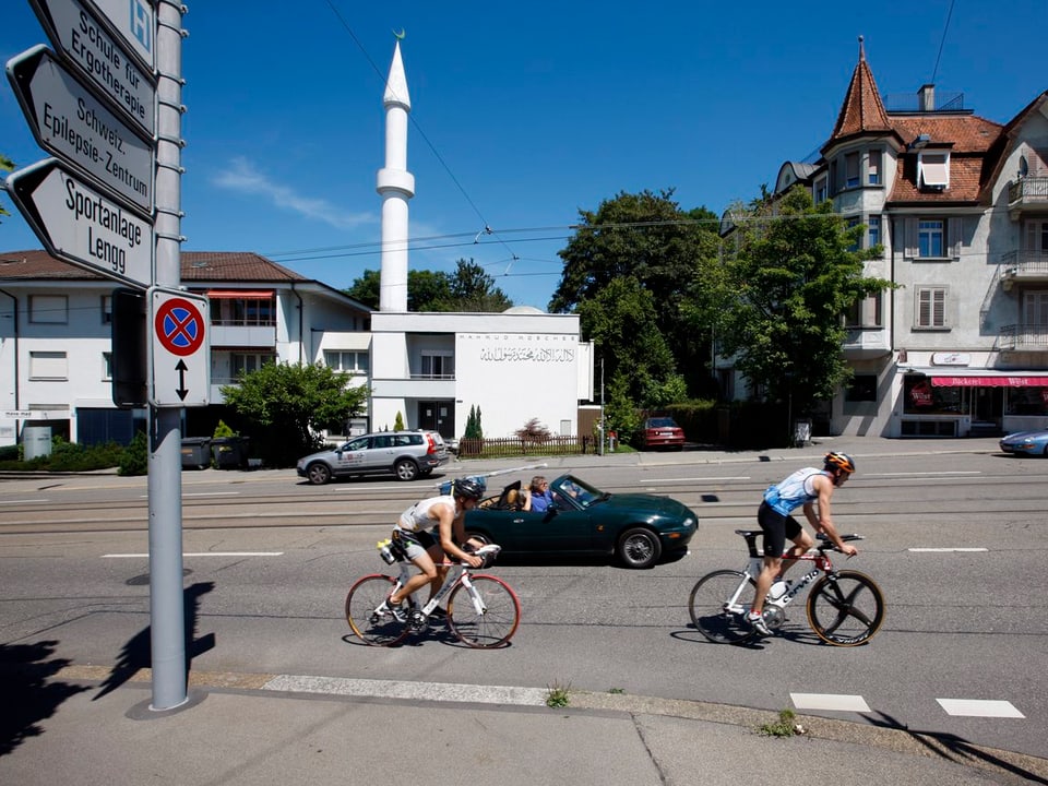 Eine weisse Moschee mit Minarett zwischen zwei Wohngebäuden an einer Strasse.