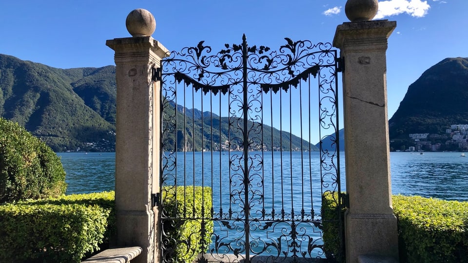 Blick durch ein Eisentor auf den blauen Luganersee.