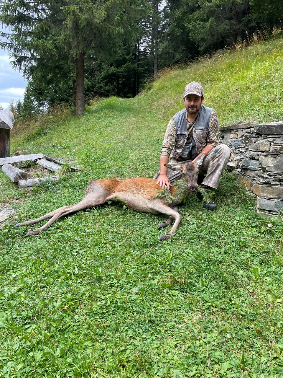 Mann in Jagdkleidung sitzt neben erlegtem Hirsch auf einer Wiese.