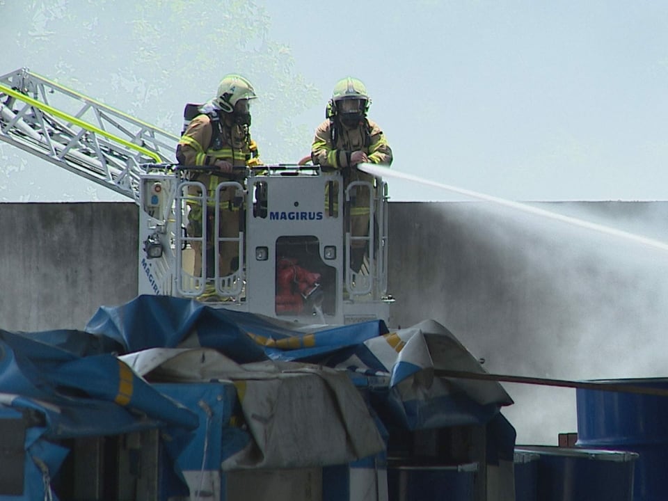 Zwei Feuerwehrleute auf einer Art Hebebühne löschen mit einem Schlauch einen Brand.