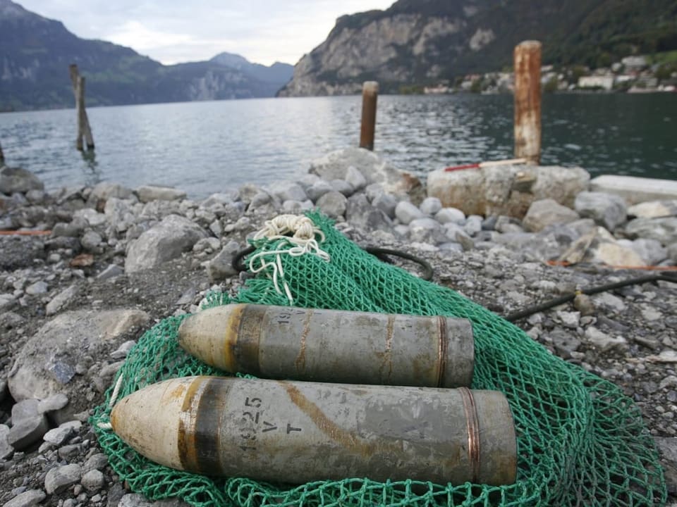 Zwei Artilleriegeschosse auf Felsstrand vor Bergsee.