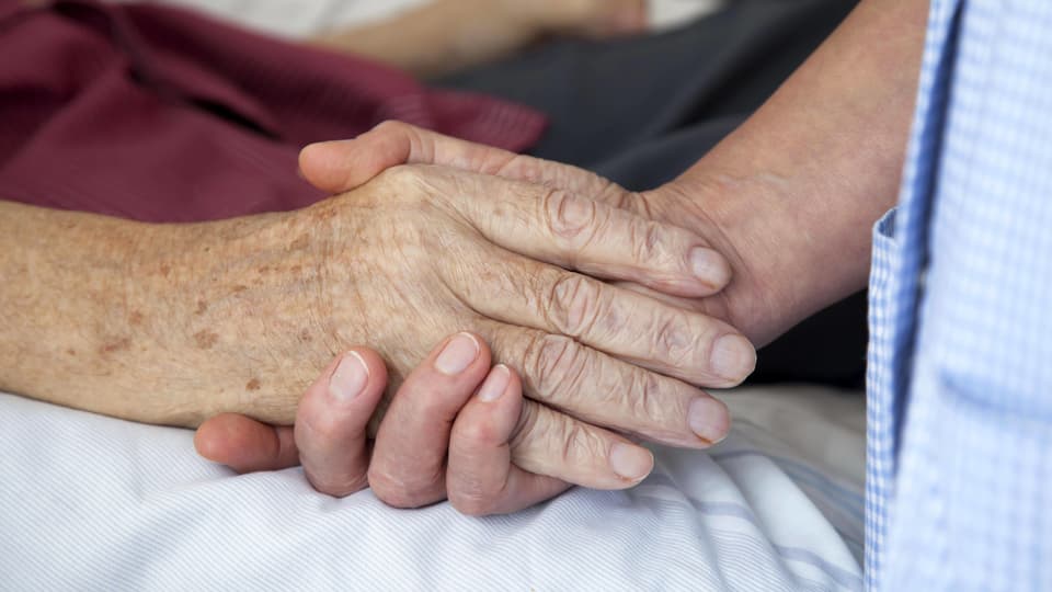 Eine Pflegende hält einem älteren Mann auf einem Bett die Hand.