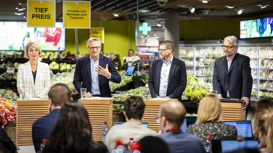 Menschen bei einer Pressekonferenz im Supermarkt, Obst- und Gemüsebereich.