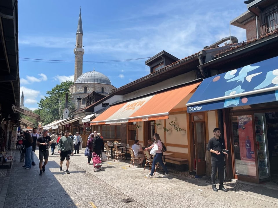 Menschen spazieren in einer Strasse mit Geschäften und einer Moschee im Hintergrund.