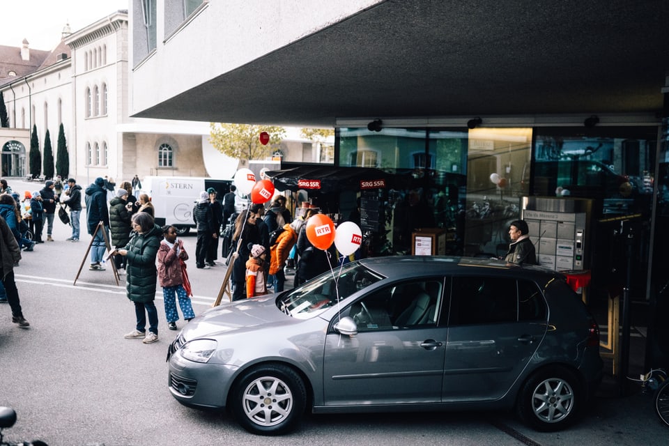 Menschengruppe vor einem Gebäude mit parkendem Auto und Luftballons.