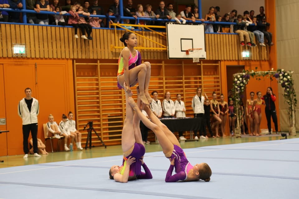 Drei Turnerinnen bei einer Akrobatik-Performance in einer Sporthalle.