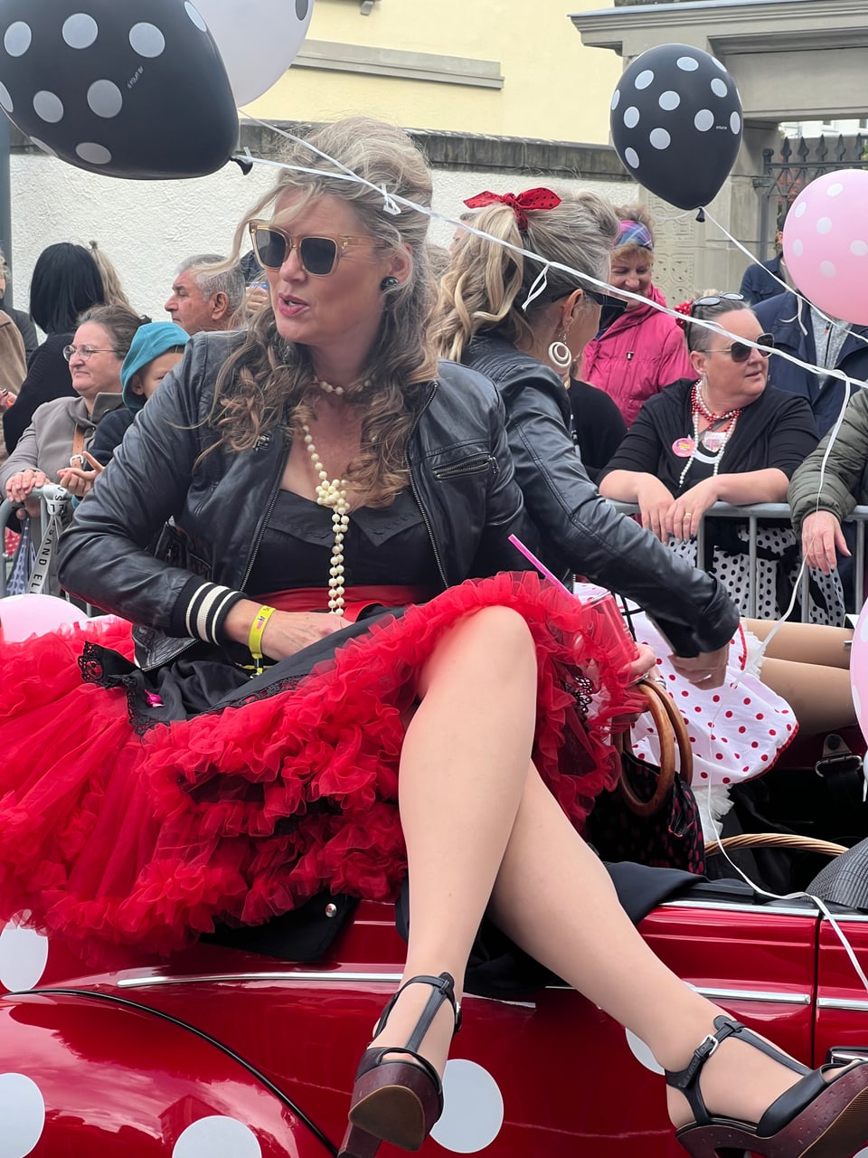 Frau in rotem Rock auf Auto bei Strassenparade mit Luftballons.