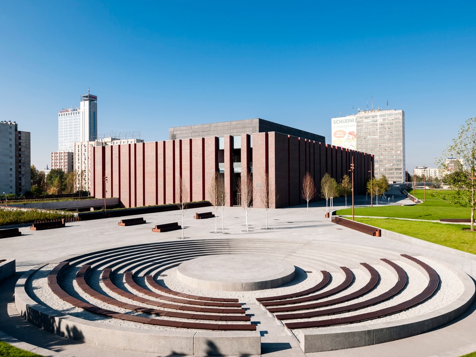 Kubistisches Gebäude aus dunkelrotem Backstein steht in einer modernen Parkanlage.