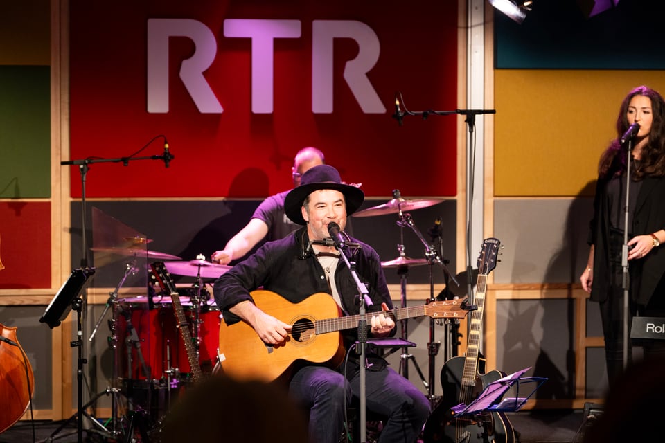 Musiker mit Gitarre vor rotem RTR-Logo auf Bühne.