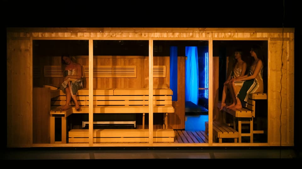 Eine Sauna, vorne offen und der Zuschauer blickt hinein. Darin sitzen drei Frauen mit Tüchern bedeckt. 