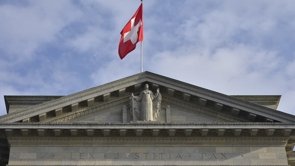 Gebäudefassade mit Schweizer Flagge und lateinischer Inschrift.