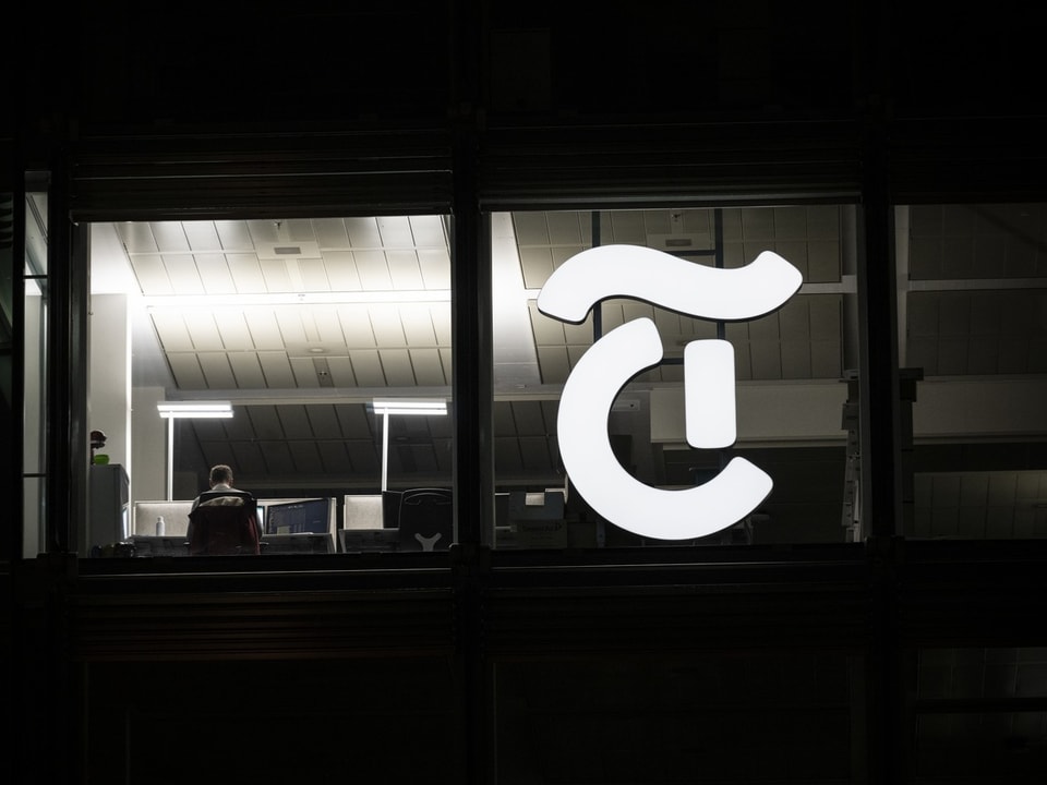 Mann in Büro mit grossem beleuchteten Logo des Buchstabens T am Fenster.