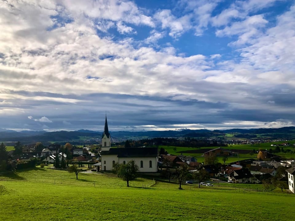 Dorfkirche im Grünen mit bewölktem Himmel und Hügeln im Hintergrund.