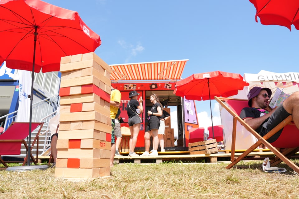 Personen auf einem Festival mit grossen Jenga-Blöcken und roten Sonnenschirmen.