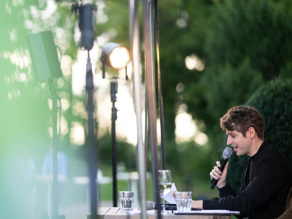 Junger Mann mit schwarzem Pullover an Tisch, Mikrofon in der Hand, im Hintergrund Bäume und Wiese, Schweinwerfer oben.