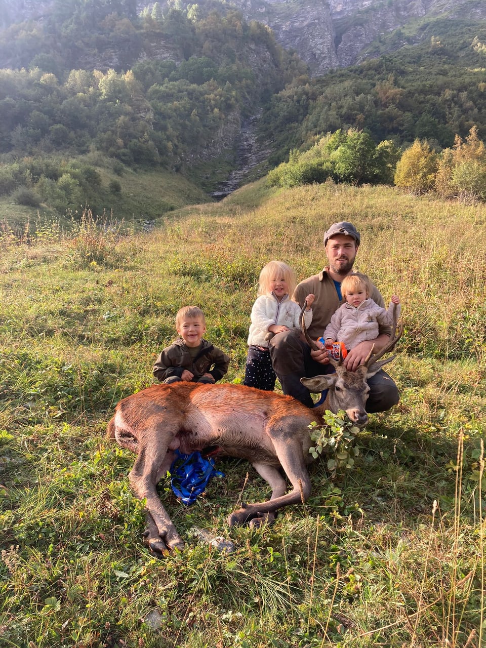 Mann mit drei Kindern neben erlegtem Hirsch im Freien.