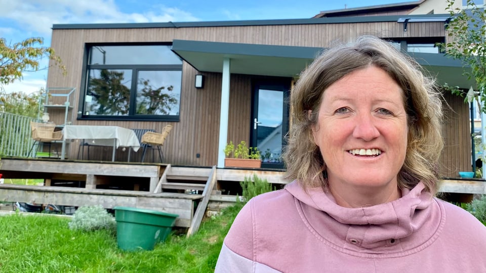 Frau in pinkem Pulli lacht vor einem kleinen Holzhaus.