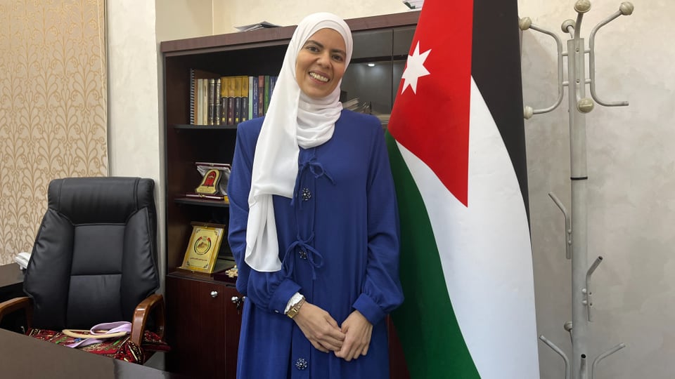 Frau in blauem Kleid steht vor jordanischer Flagge im Büro.