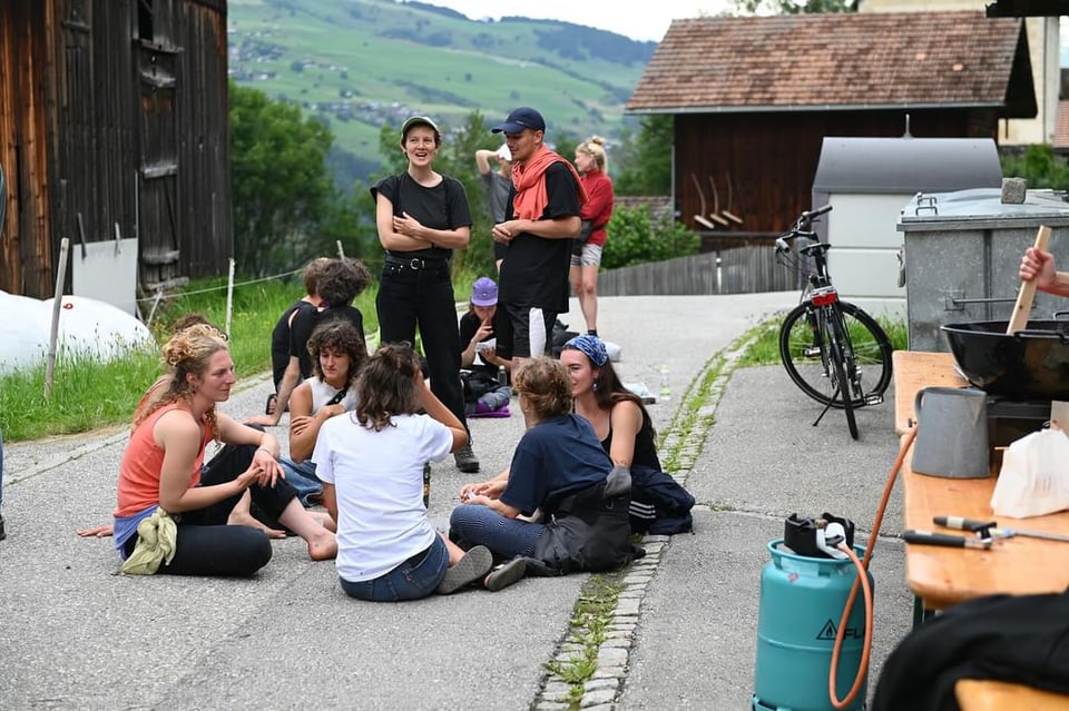 Gruppe von Menschen sitzt und steht auf einer Strasse in einer ländlichen Umgebung.