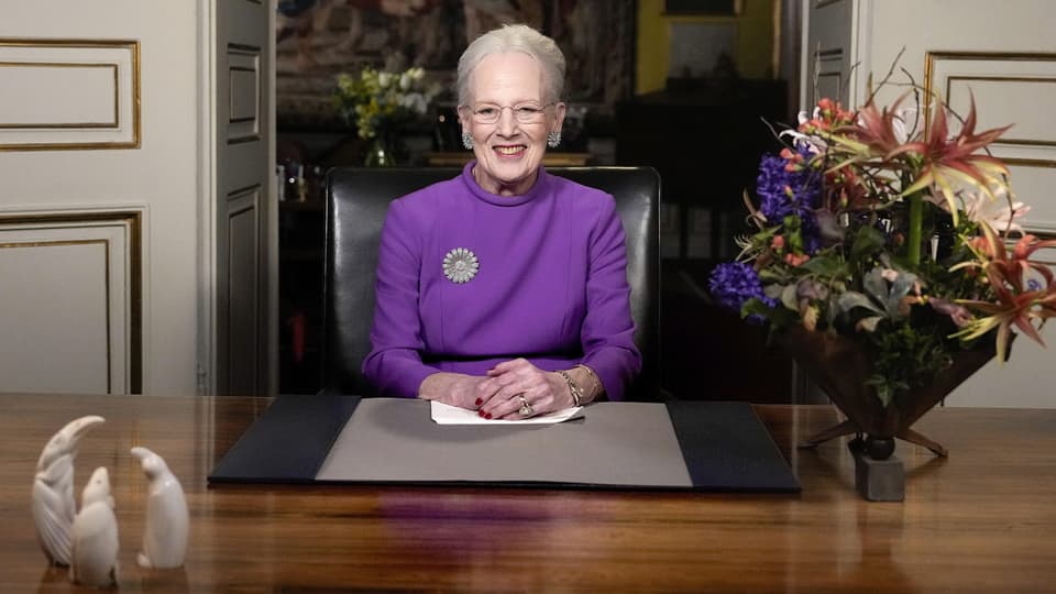 Margrethe II. vor einem Pult. Rechts von ihr befindet sich ein Blumenstrauss, sie trägt ein violettes Kleid.