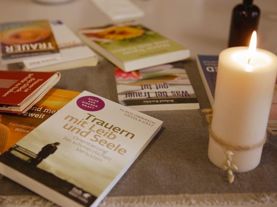 Eine weisse Kerze brennt auf einem Tisch. Daneben liegen verschiedene Selbsthilfebücher für Hinterbliebene.
