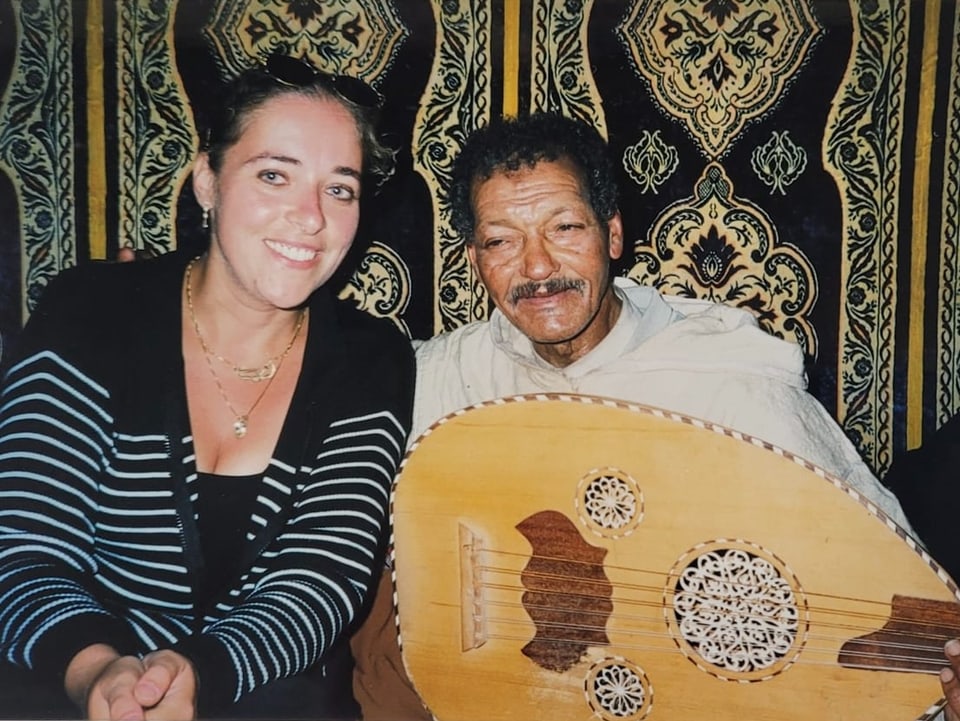 Eine Frau mit dunklem Haar lächelt in die Kamera. Neben ihr sitzt ein älterer Mann mit gitarrenähnlichem Instrument.