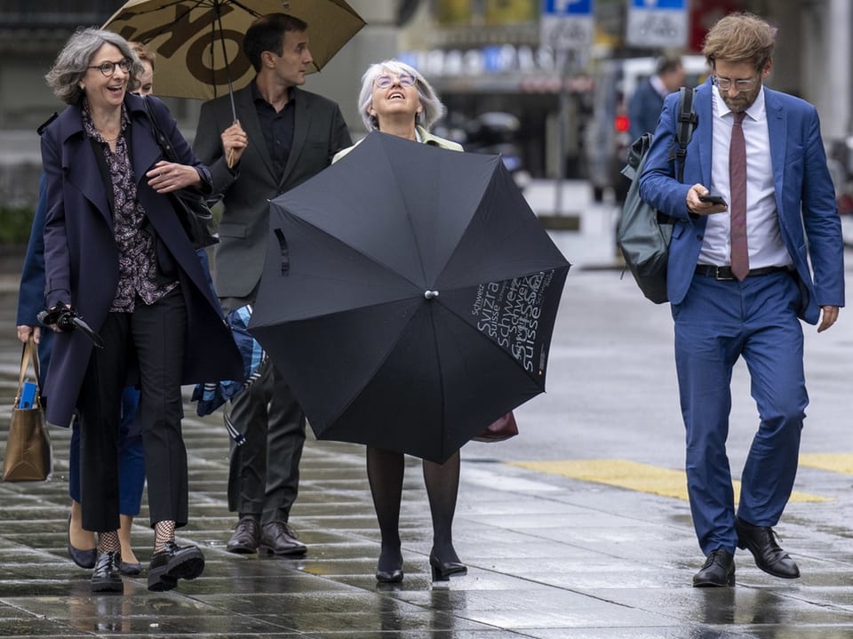 Vier Personen gehen im Regen auf dem Bürgersteig; eine Frau hält schwarzen Schirm.