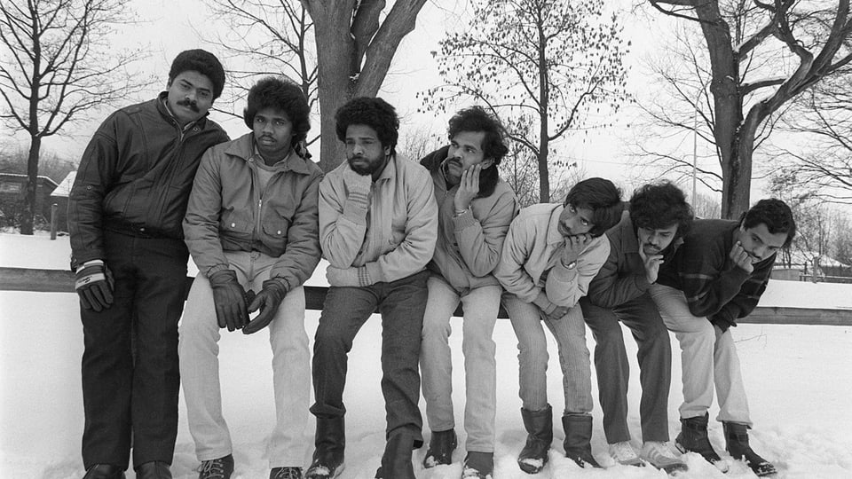 Sieben Männer lehnen sich sitzend an Reling an; im Hintergrund viel Schnee