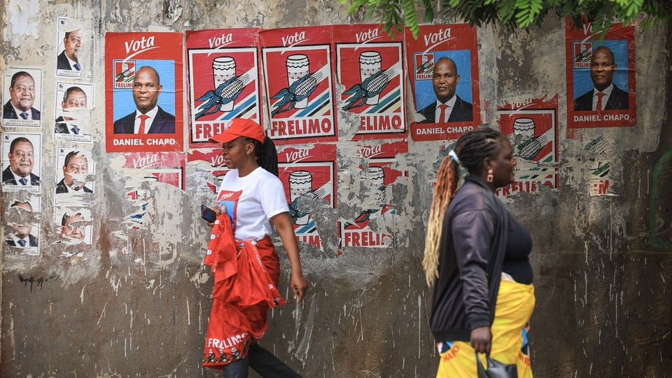 Flyer von politischen Gruppen hängen an der Wand, in den roten Farben der FRELIMO. Zwei Frauen laufen daran vorbei. 