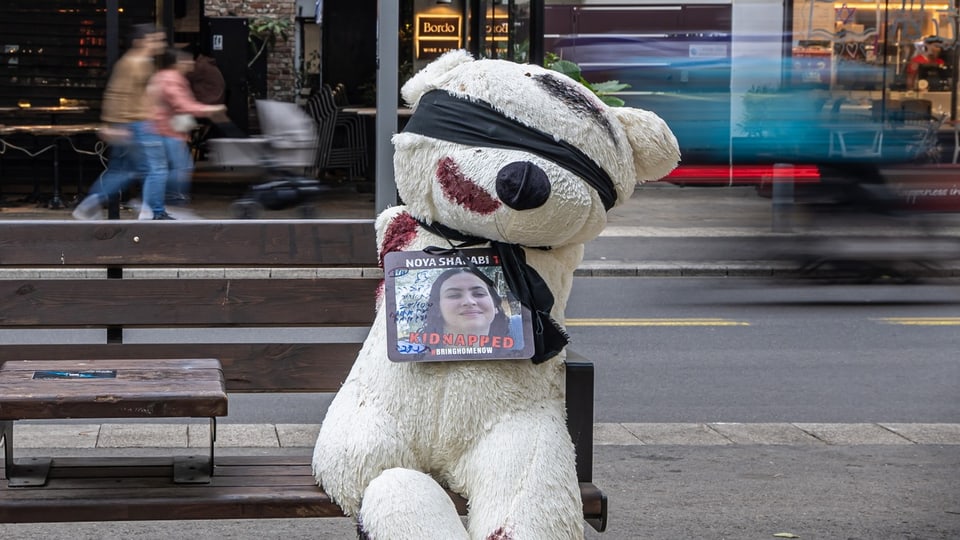 Ein Teddybär mit verbundenen Augen, der das Bild eines von Hamas-Terroristen entführten Mädchens hält, auf einer Bank
