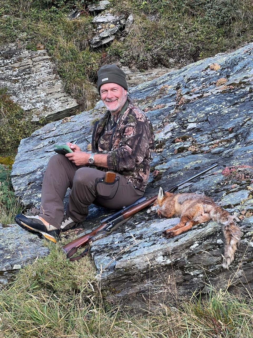 Mann im Tarnanzug sitzt auf Felsen, Gewehr und erlegter Fuchs neben ihm.