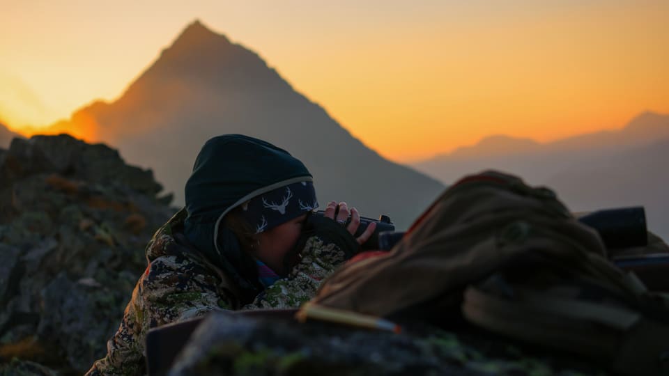 Vermummte Person beobachtet durch Fernglas mit Berg im Sonnenuntergang.