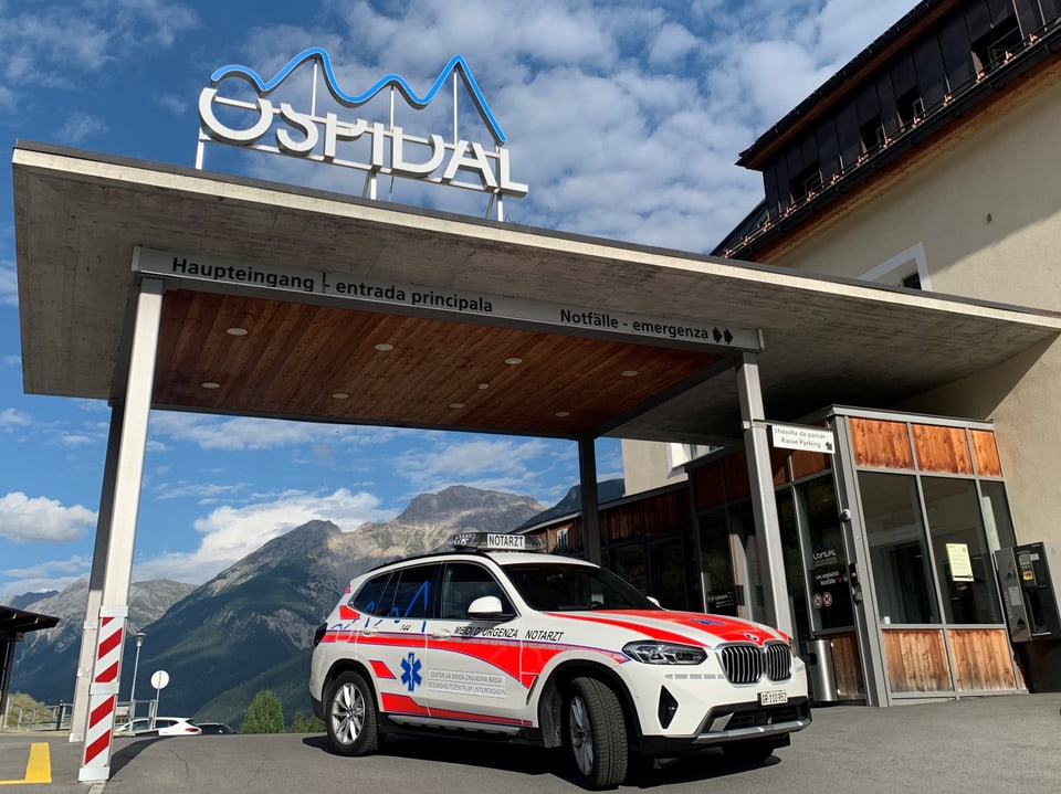 Ein Krankenwagen vor dem Eingang eines Krankenhauses in alpiner Umgebung.