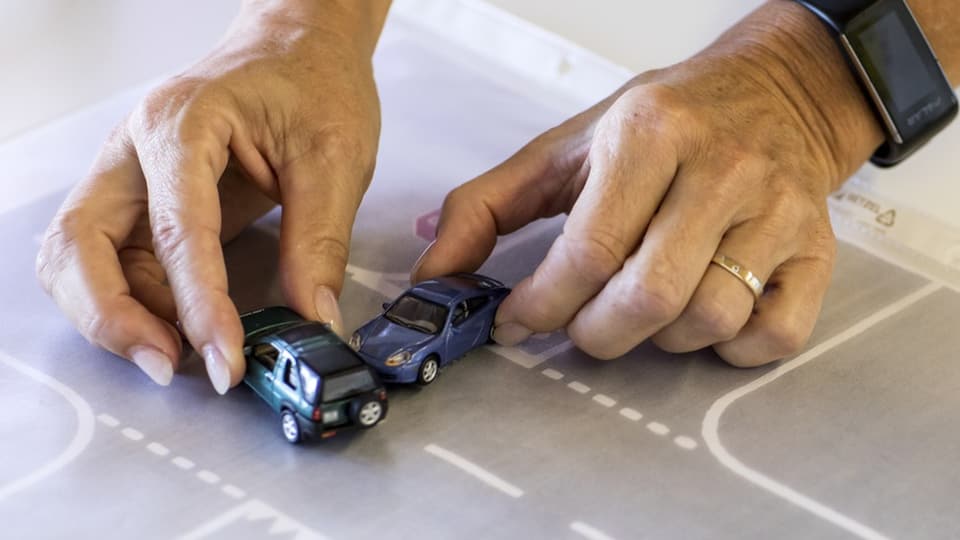 Zwei Hände steuern auf einem Papier mit eingezeichneter Strasse zwei Spielzeugautos.