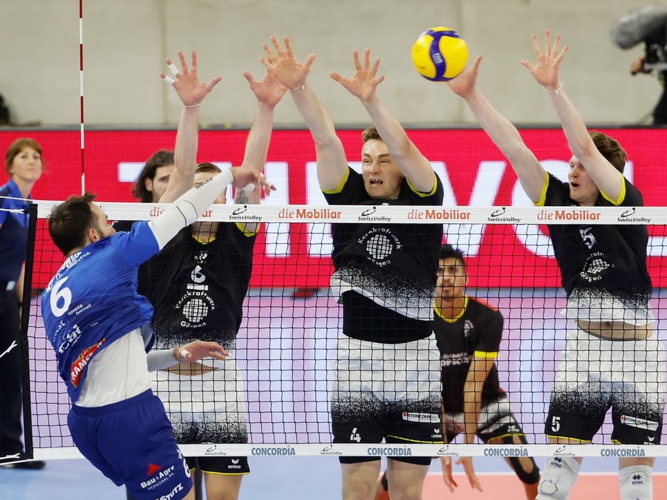 Volleyballspiel mit Spieler im blauen Trikot gegen drei Spieler im schwarzen Trikot am Netz.