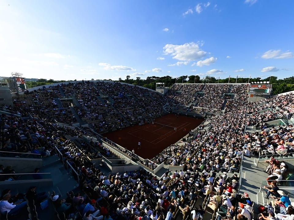 Grosses Tennisstadion mit vielen Zuschauern und blauem Himmel.