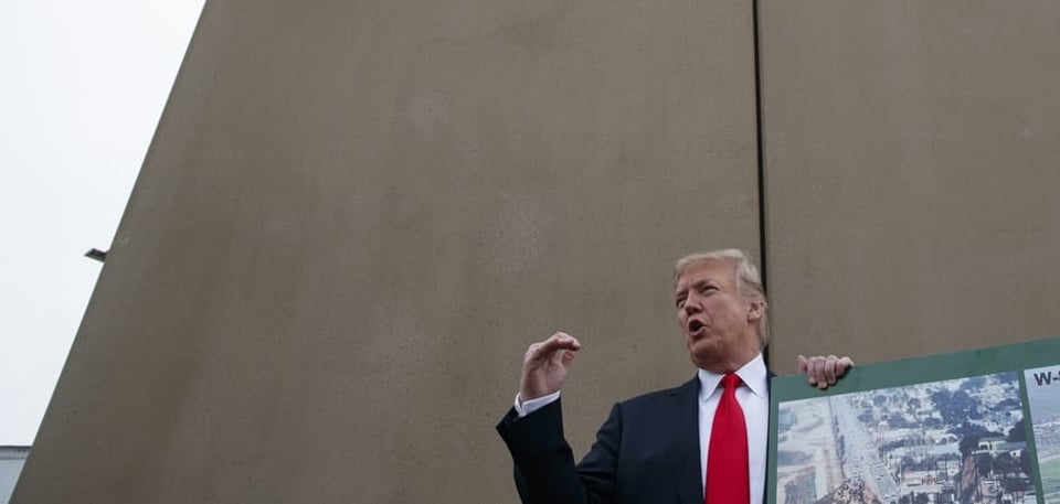 Donald Trump vor einer hohen grauen Betonmauer.