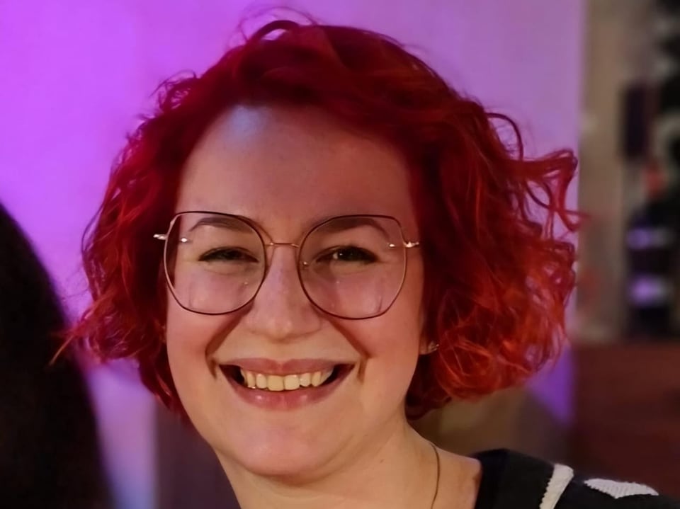 Frau mit rotem lockigem Haar und Brille lächelt.