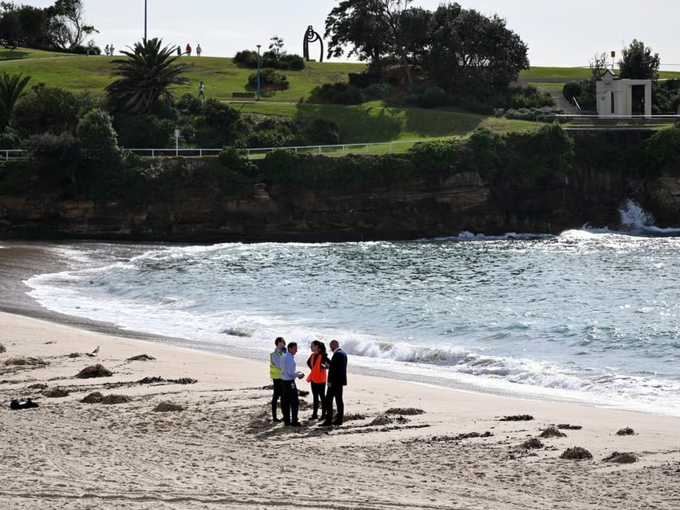 Menschen stehen am Strand vor einer felsigen Küste.