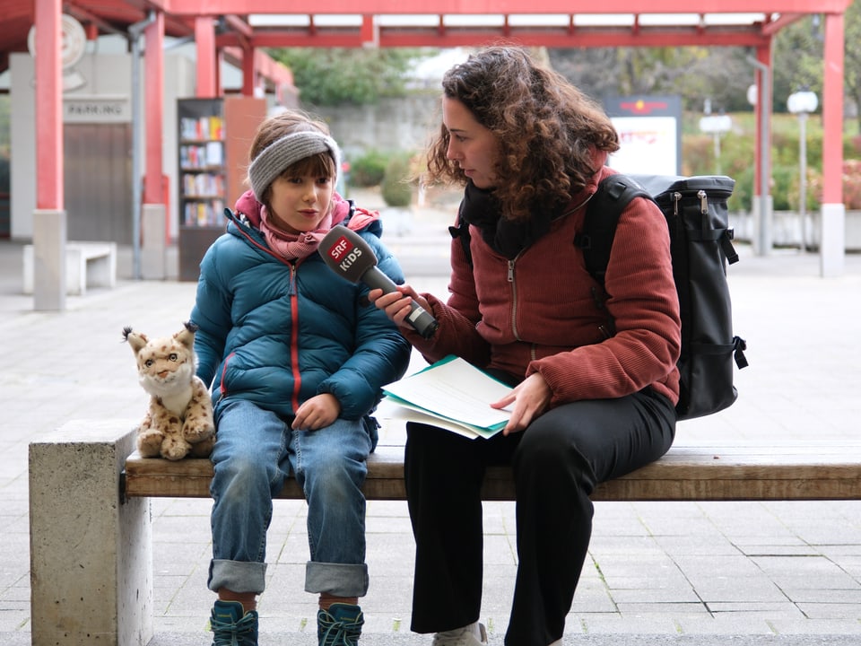 Moderatorin Luana sitzt mit Kinderreporterin Ava auf einer Bank. Neben ihnen sitzt ein kleiner Stoffluchs. 